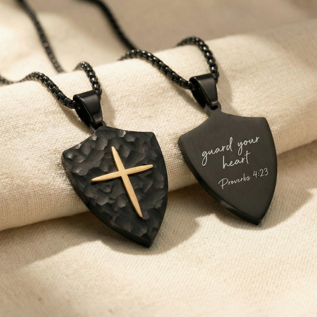 Two black shield-shaped pendants with a gold cross on a beige fabric background