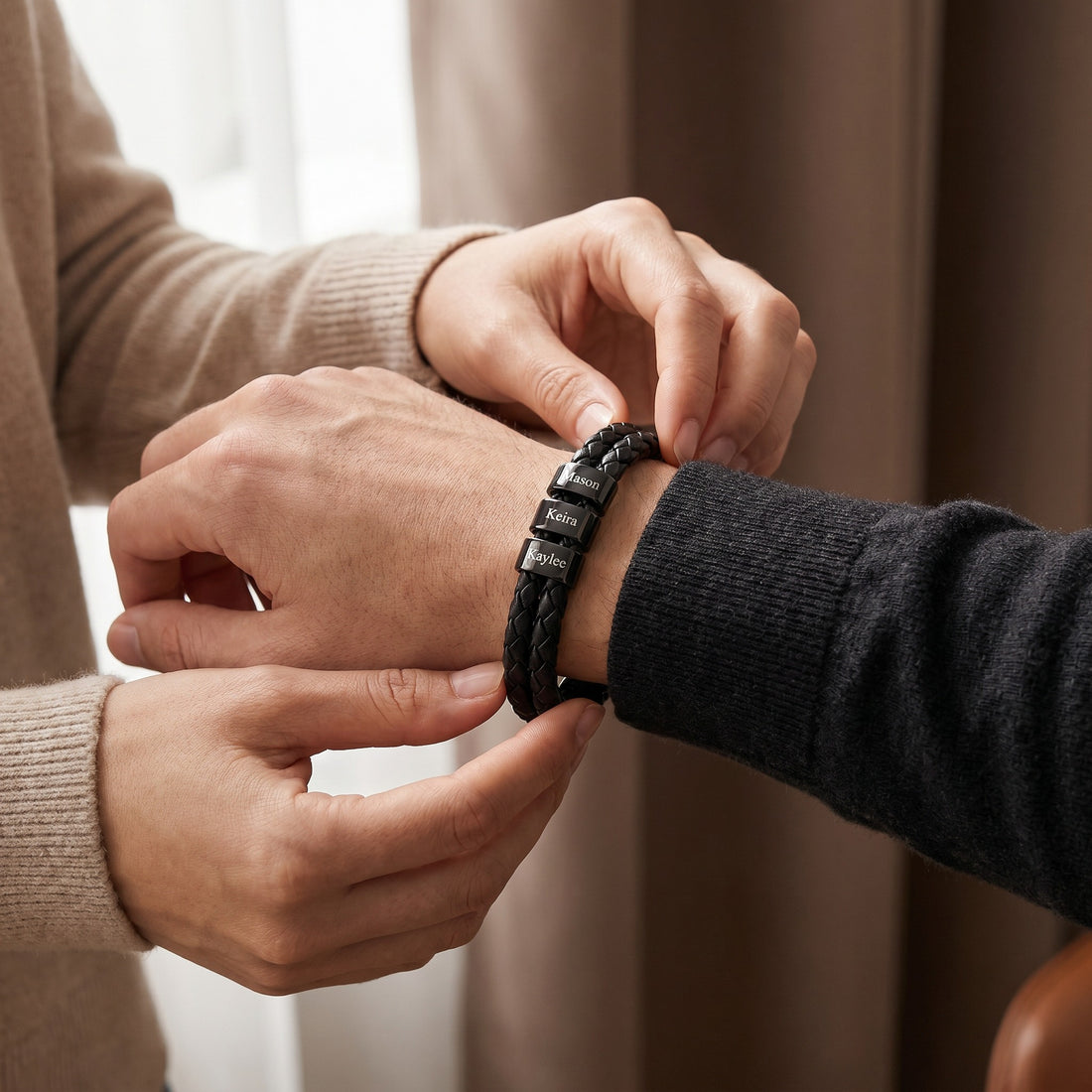 Two people adjusting a black braided bracelet on a neutral background