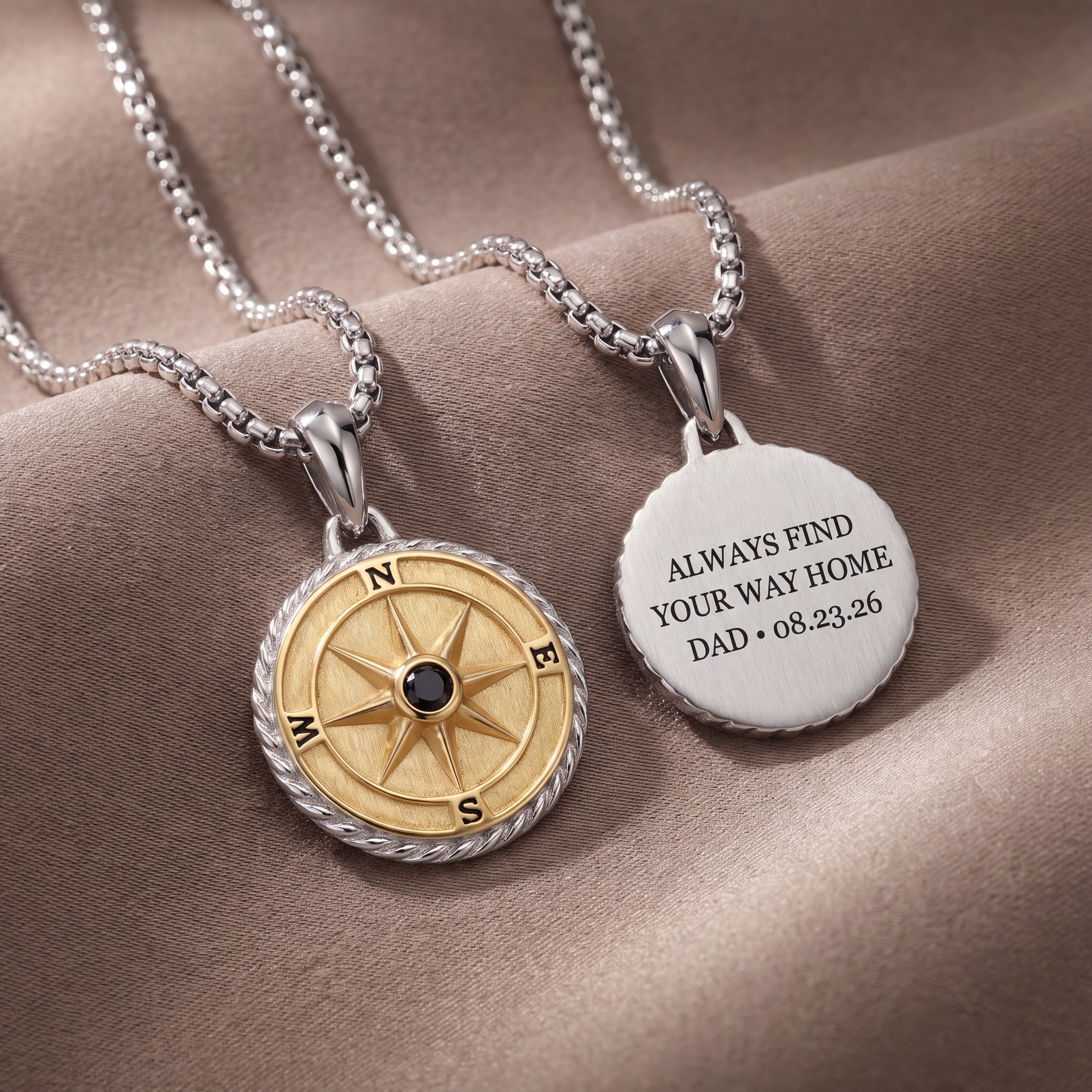 Two personalized necklaces displayed on a draped brown fabric background. One necklace features a gold and silver compass pendant with a small black center, and the other features a round silver pendant engraved with 'ALWAYS FIND YOUR WAY HOME DAD + date
