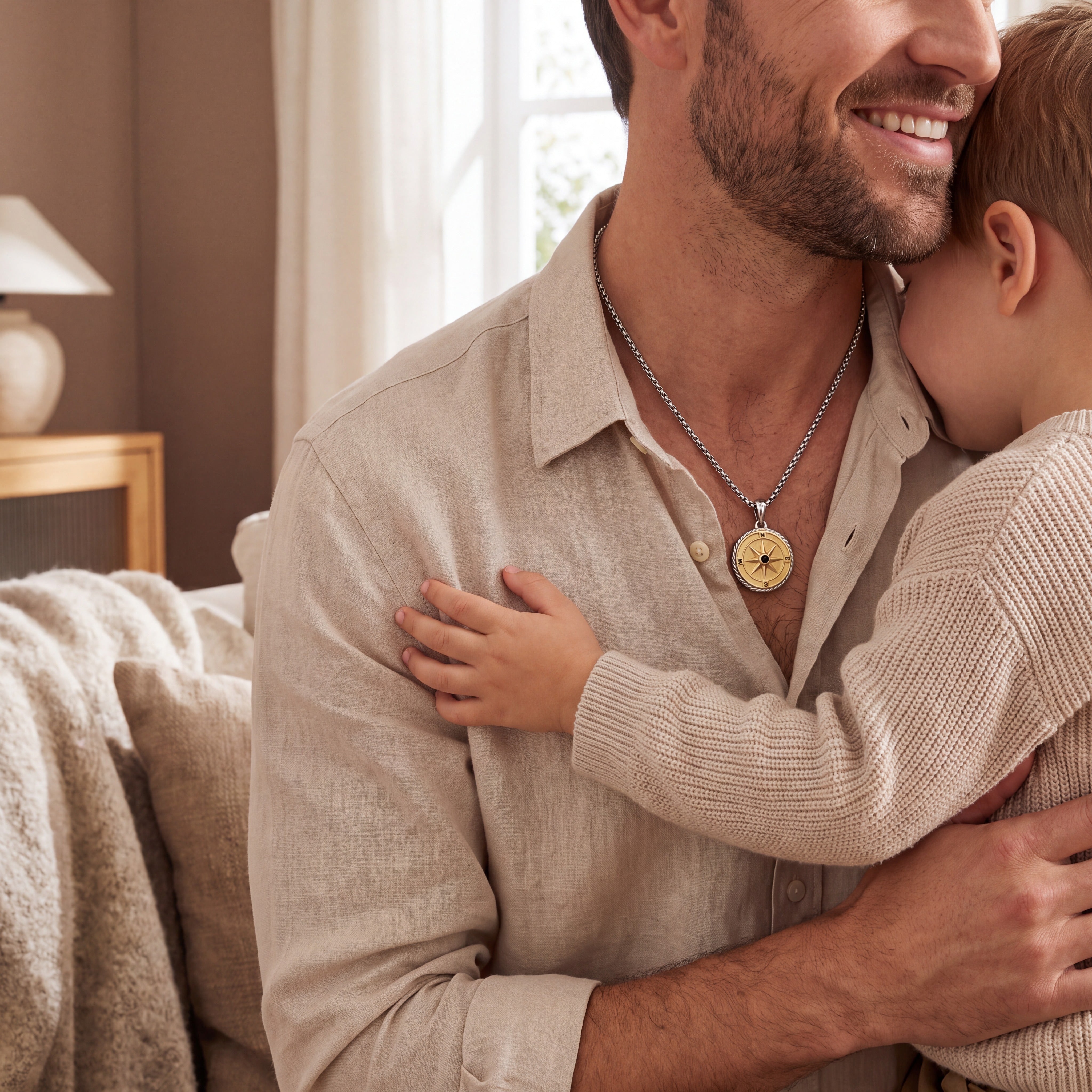 A man and a child hugging indoors, with the man wearing a silver chain necklace featuring a gold-toned compass pendant. The scene is softly lit with warm neutral tones and a cozy home background.