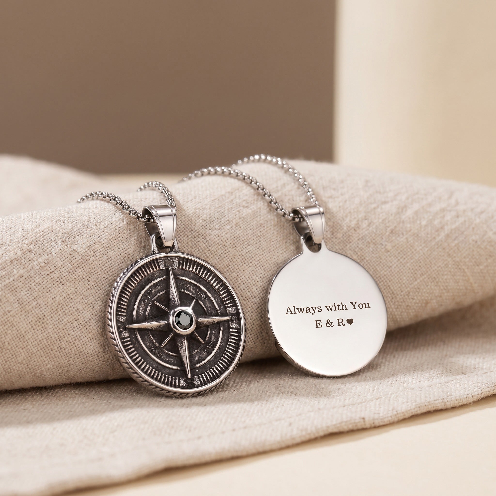Two silver necklaces with a compass pendant and engraved round pendant on beige fabric.