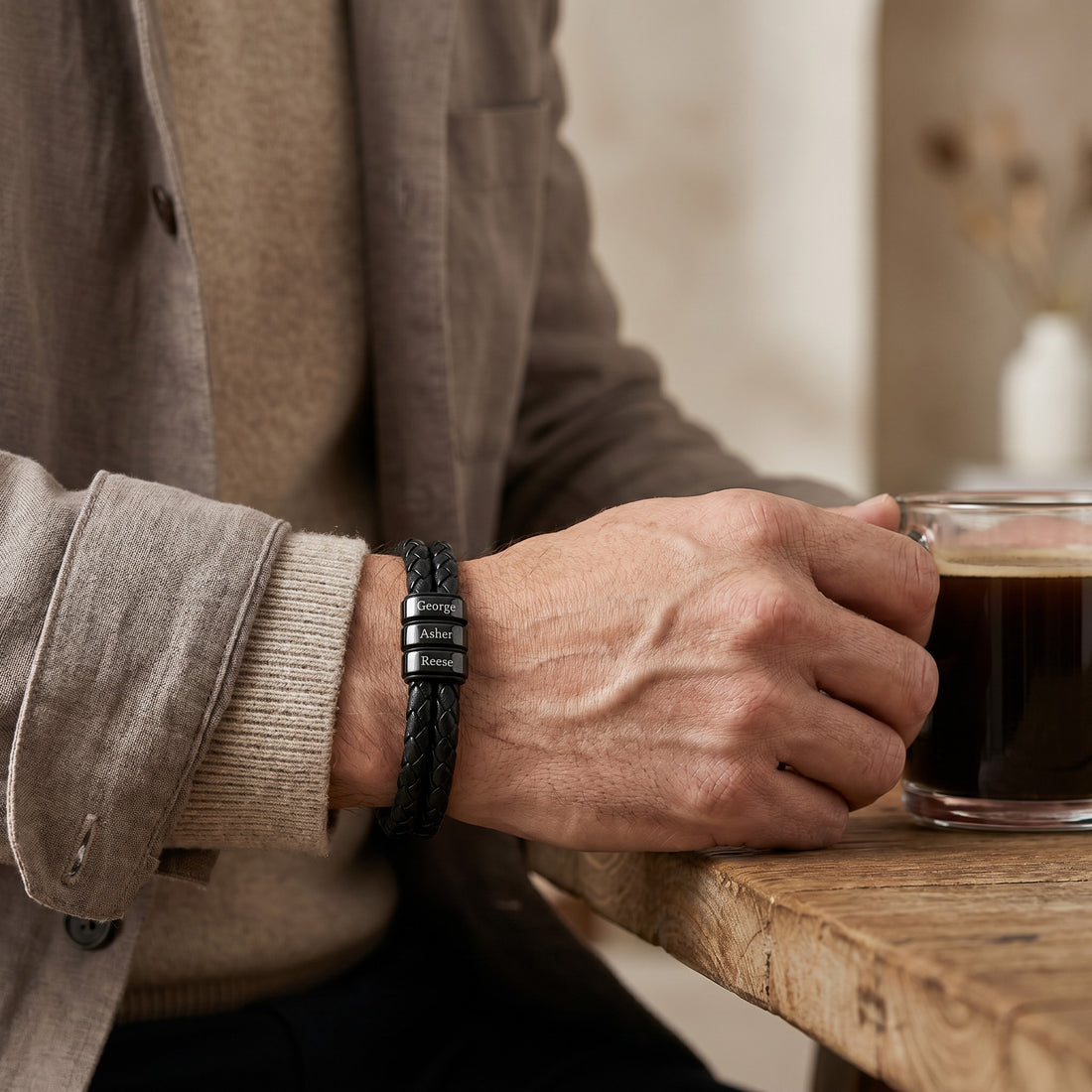 Person wearing a leather bracelet holding a glass of dark liquid, possibly coffee, on a wooden surface.