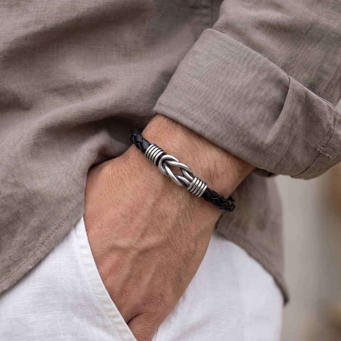 A close-up of a man's wrist wearing a black braided leather bracelet with a silver-toned metal knot closure, styled with a taupe linen shirt and white trousers.
