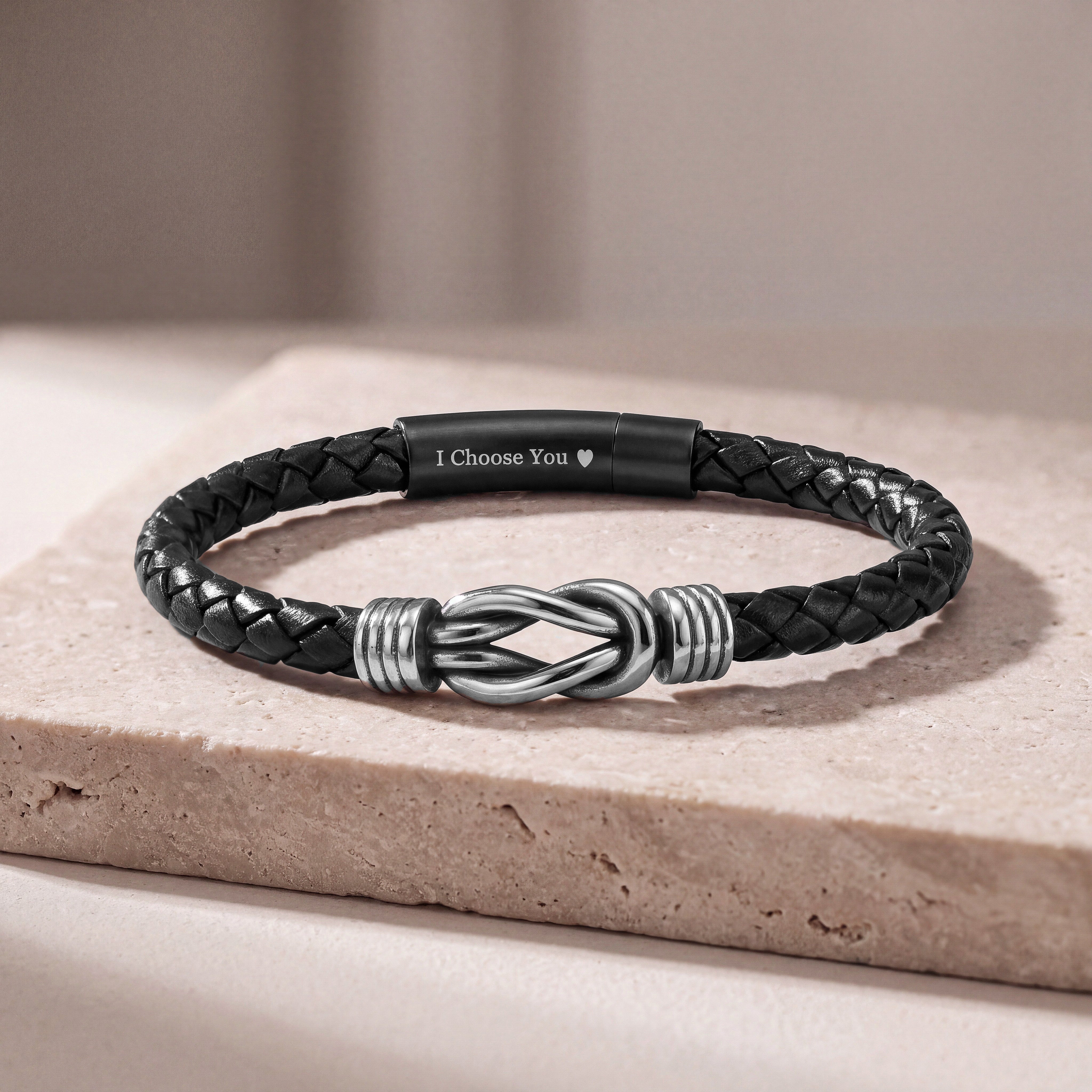 A black braided leather bracelet with a silver infinity knot centerpiece and a black magnetic clasp engraved with 'I Choose You', resting on a beige stone slab.