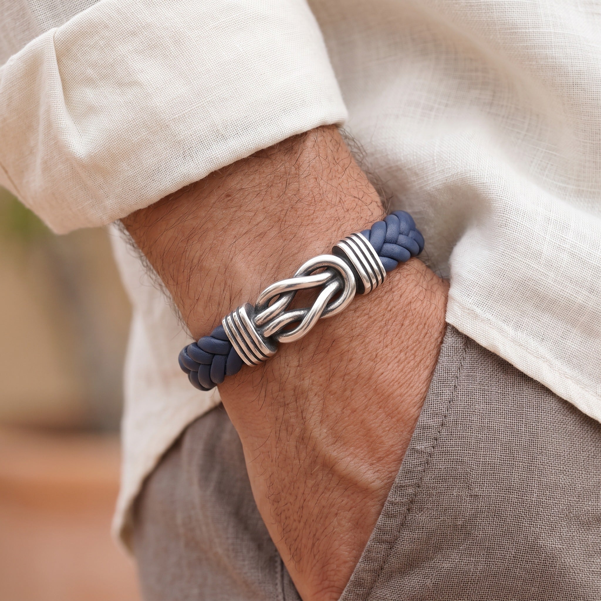 A close-up of a man's wrist wearing a braided navy blue and silver metal bracelet, styled with a cream linen shirt and gray trousers.