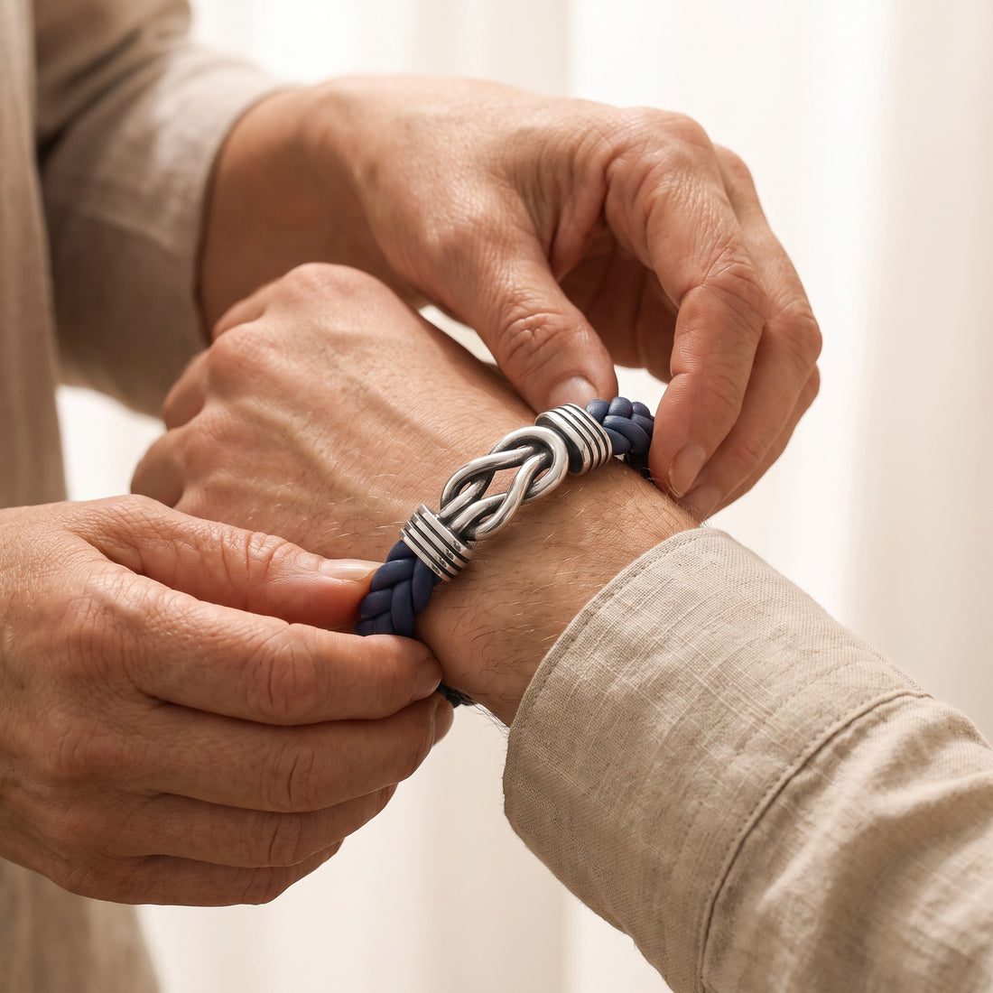 A close-up of a person's hands adjusting a braided navy blue and silver metal bracelet on another person's wrist, set against a soft, light beige background.