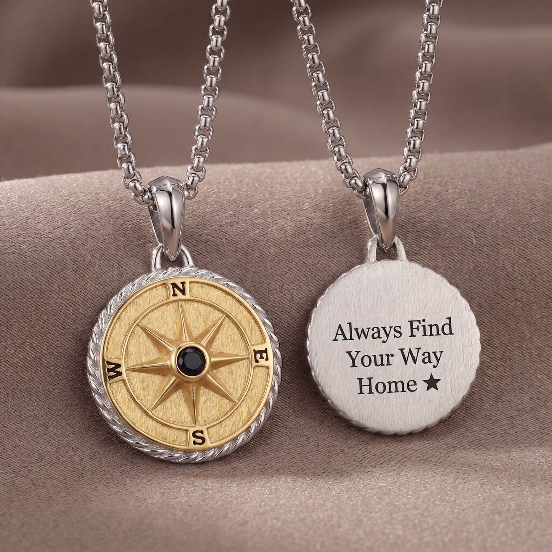 Two silver necklace pendants displayed on a soft beige fabric background. One pendant is a round compass design with a gold-toned center, black directional letters, and a small black stone at the center.