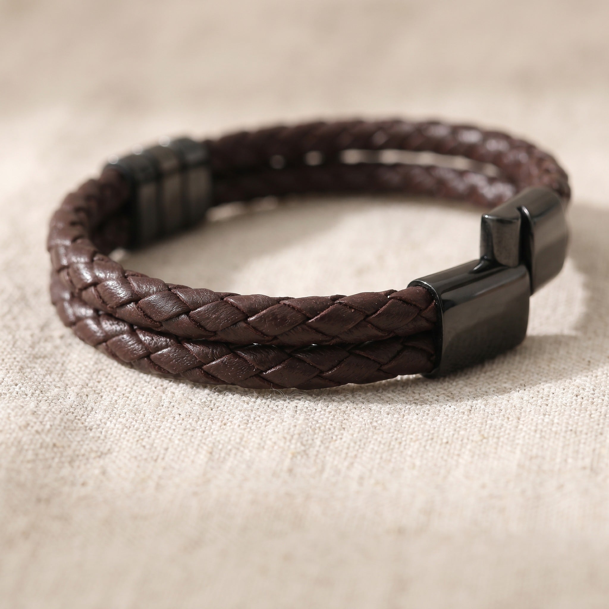 Brown braided leather bracelet with a black clasp on a beige fabric background
