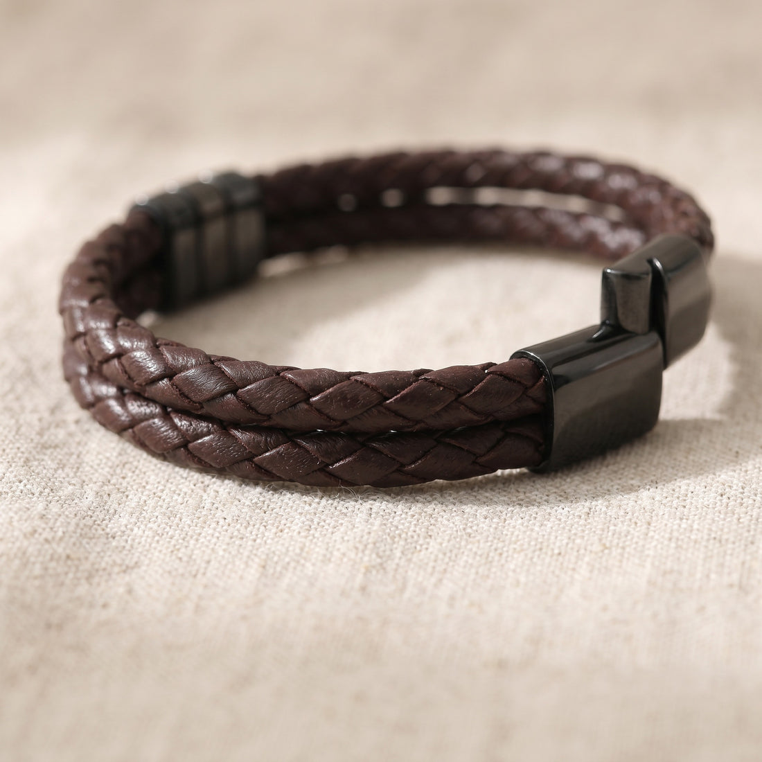 Brown braided leather bracelet with a black clasp on a beige fabric background
