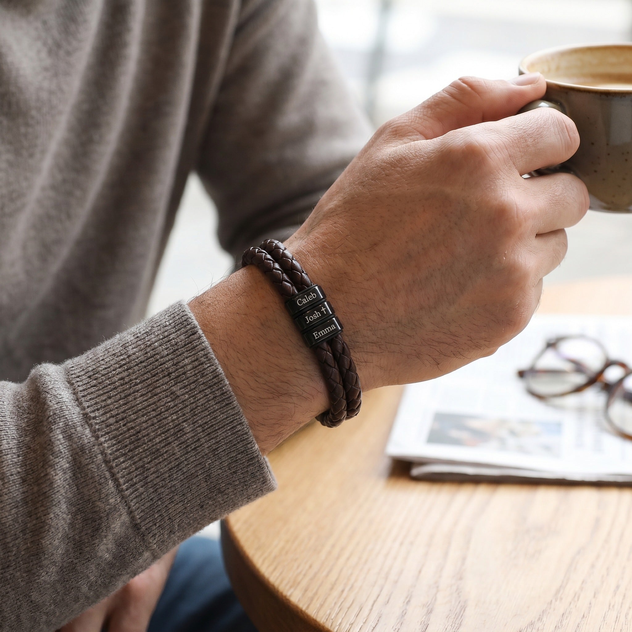 Person holding a mug with a bracelet on wrist, sitting at a table with a magazine and glasses.