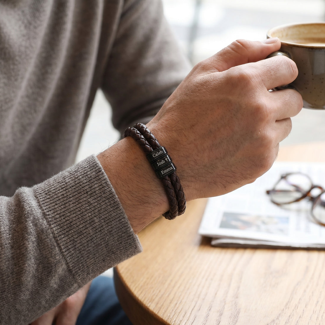 Person holding a mug with a bracelet on wrist, sitting at a table with a magazine and glasses.