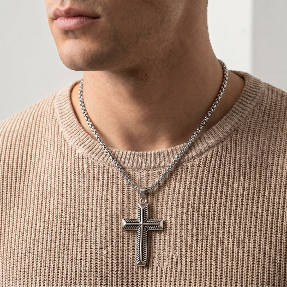 Man wearing a beige sweater with a silver cross necklace against a neutral background