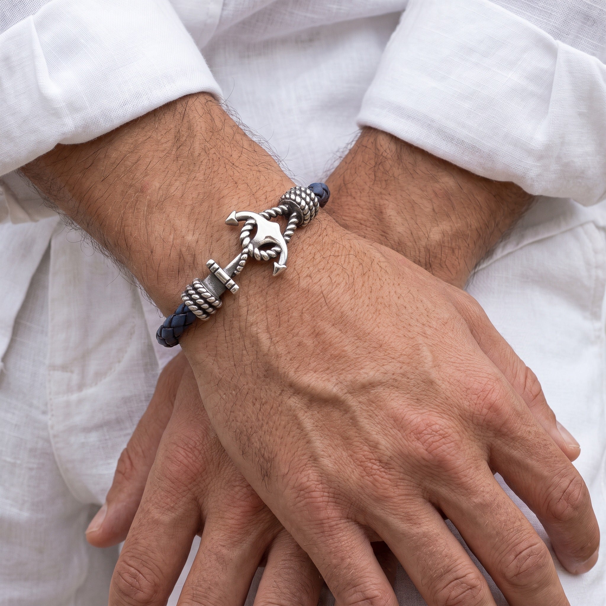 A close-up of a man's crossed hands and wrists wearing a silver-toned horse-shaped rope bracelet with dark blue cord accents, set against a white linen shirt background.