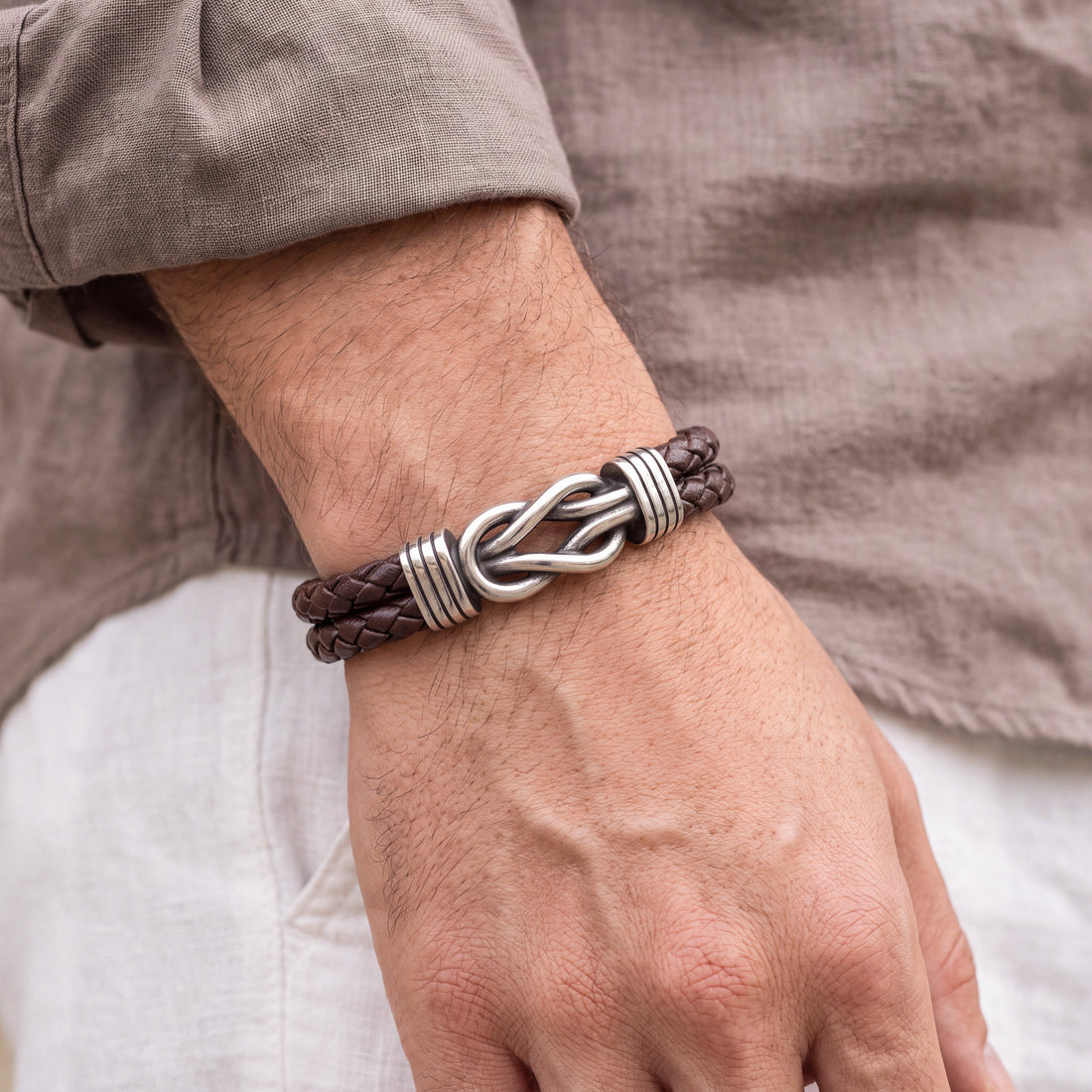 A close-up of a man's wrist wearing a dark brown braided leather bracelet with a polished silver infinity link centerpiece and textured silver end caps.