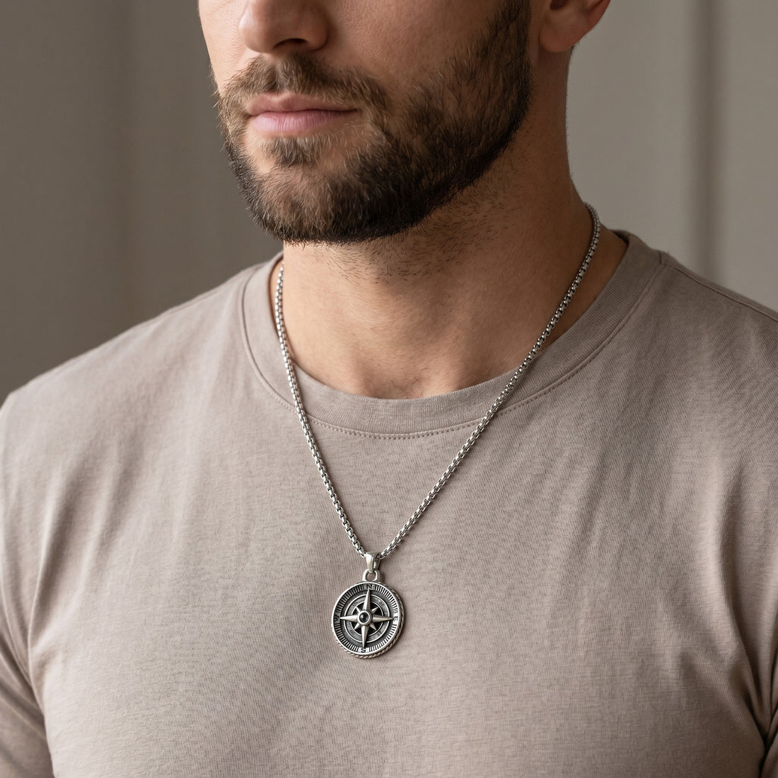 Man wearing a silver necklace with a compass pendant against a neutral background