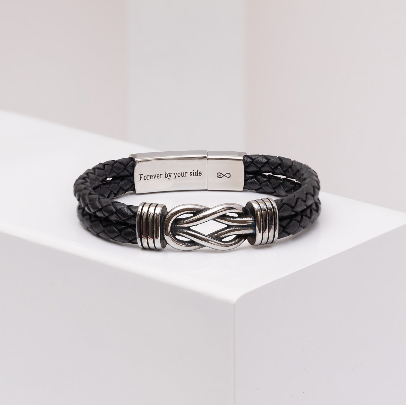 A black braided leather bracelet with a silver-toned metal clasp engraved with 'Forever by your side' and an infinity symbol, displayed on a white geometric block.