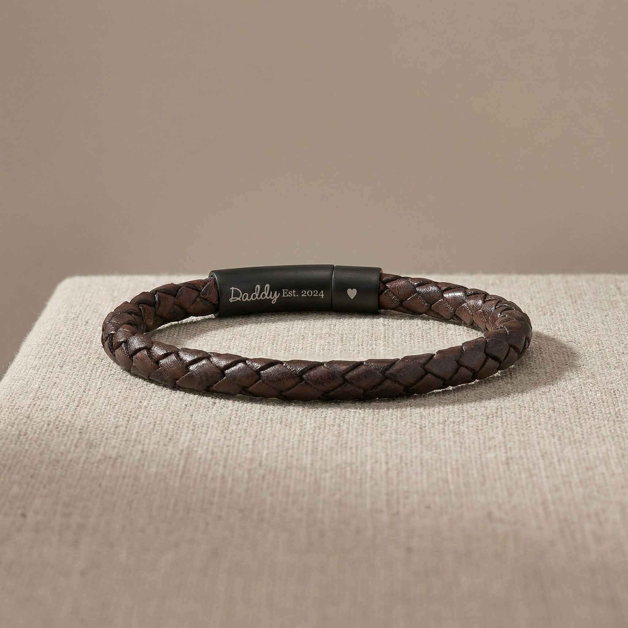 Brown braided leather bracelet with engraved tag on a beige surface