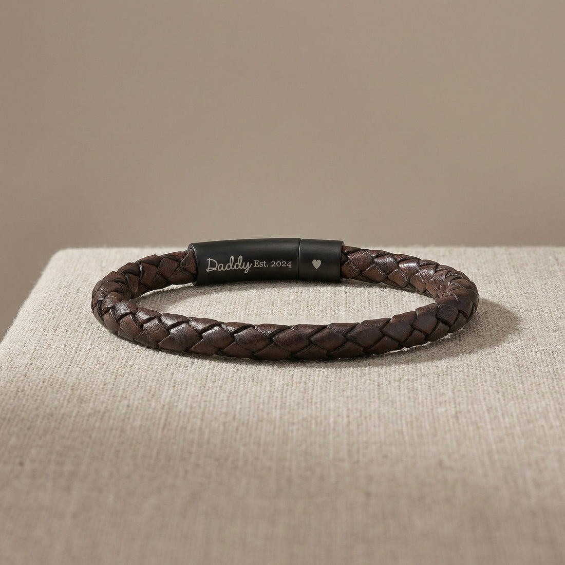 Brown braided leather bracelet with engraved tag on a beige surface