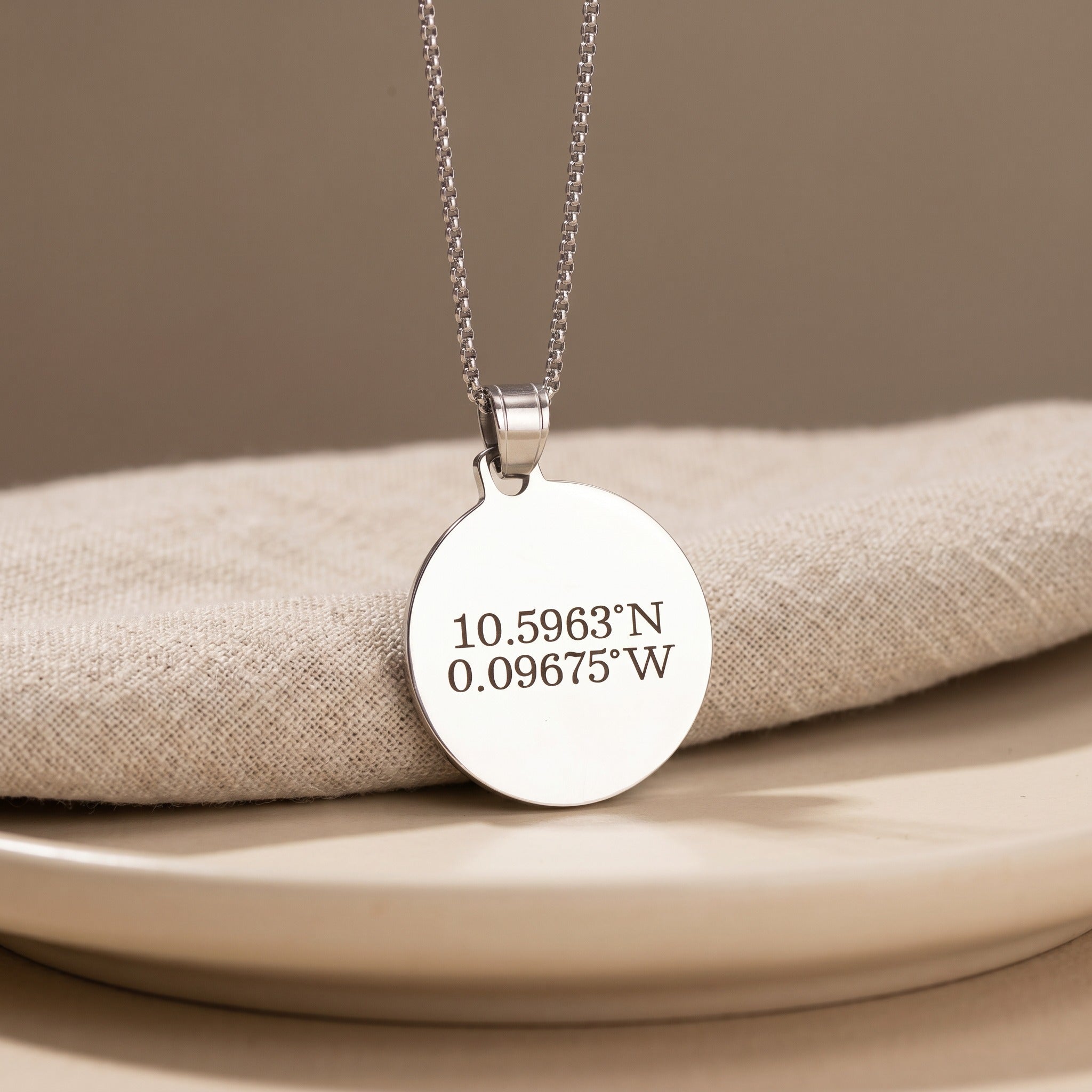 Silver necklace with a round pendant displaying coordinates on a beige surface.
