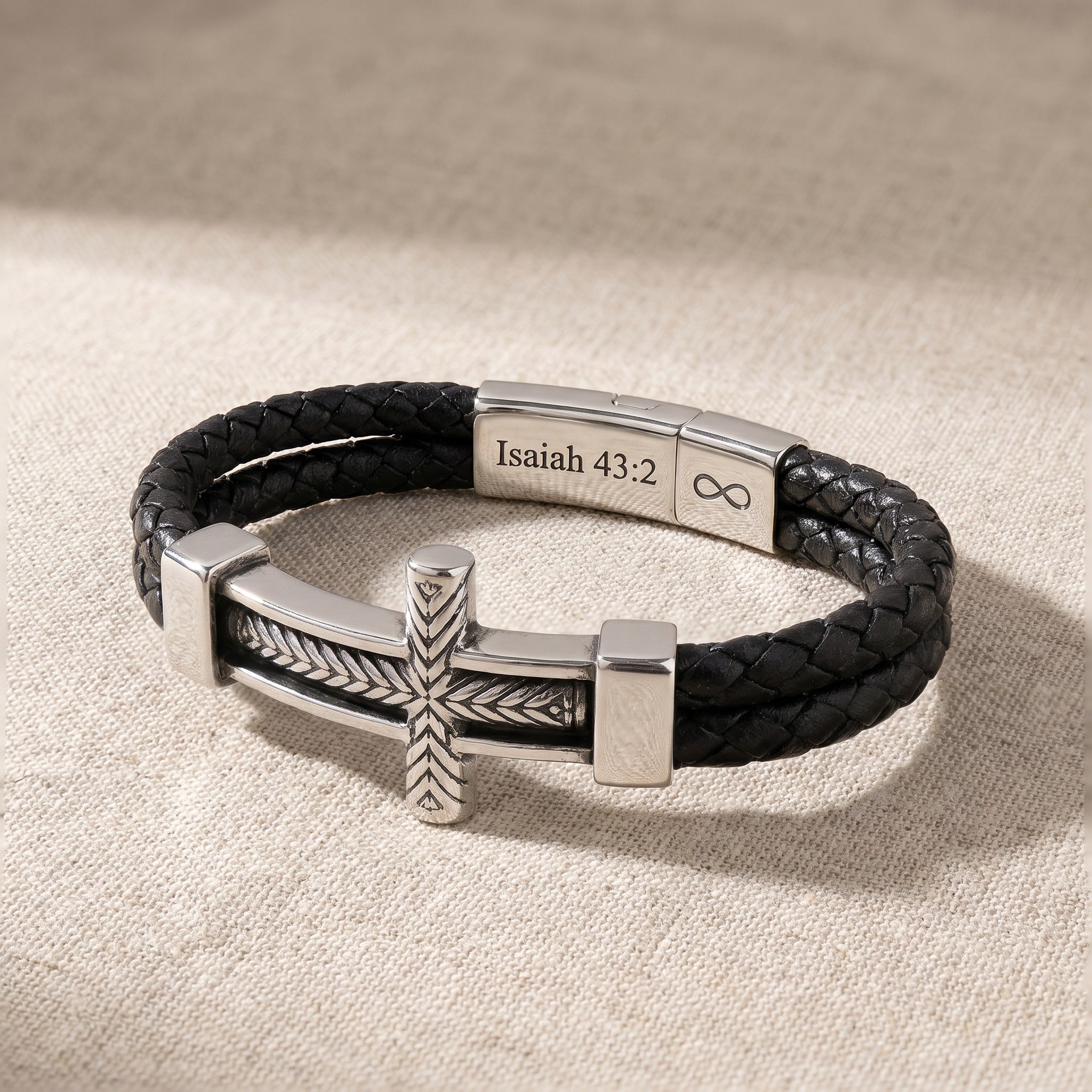 Black braided leather bracelet with silver cross and engraved bar on a beige fabric background