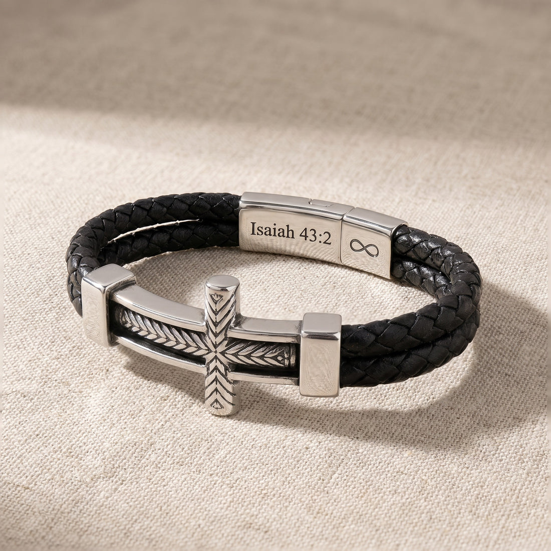 Black braided leather bracelet with silver cross and engraved bar on a beige fabric background