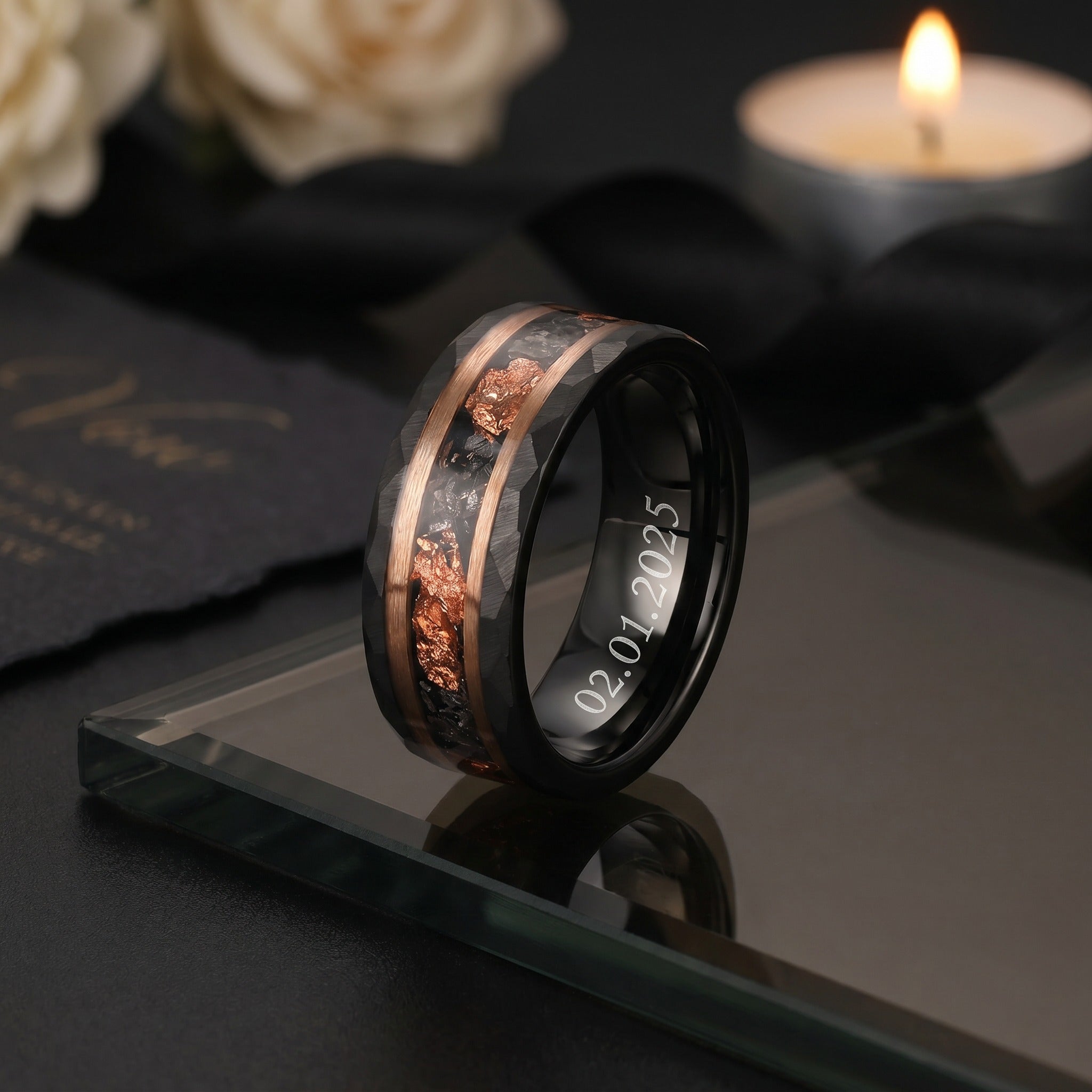 Two black wedding bands with copper inlay on a reflective surface, with a candle and flowers in the background.