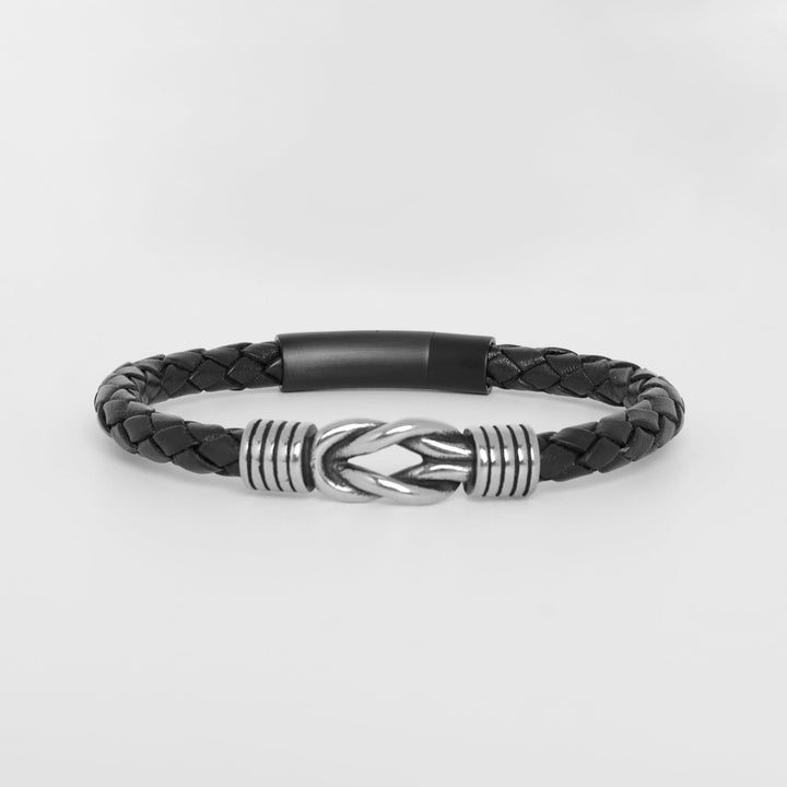 A black braided leather bracelet with a polished silver-toned metal central clasp and textured silver end spacers, displayed on a light gray background.