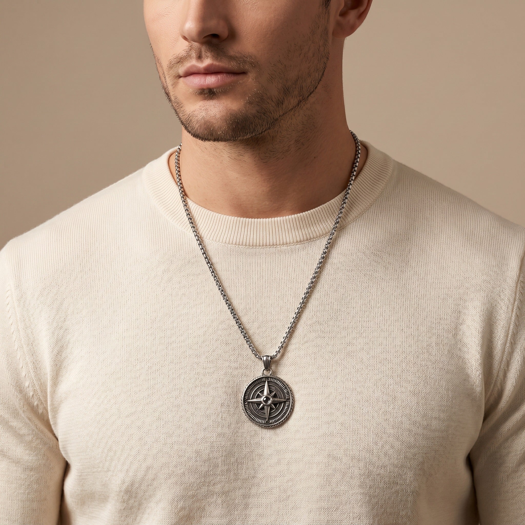 Man wearing a beige sweater with a silver necklace featuring a compass pendant against a beige background
