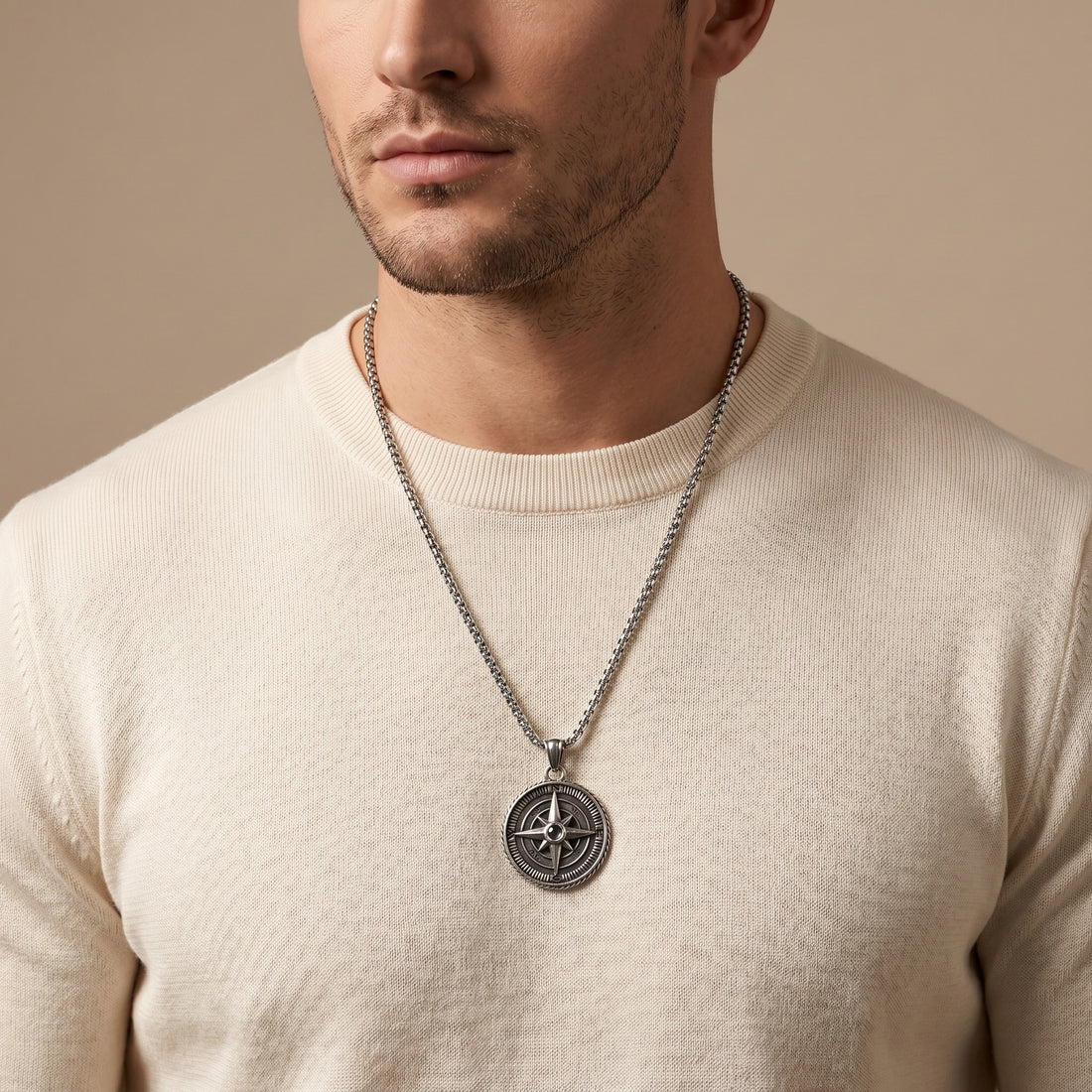 Man wearing a beige sweater with a silver necklace featuring a compass pendant against a beige background