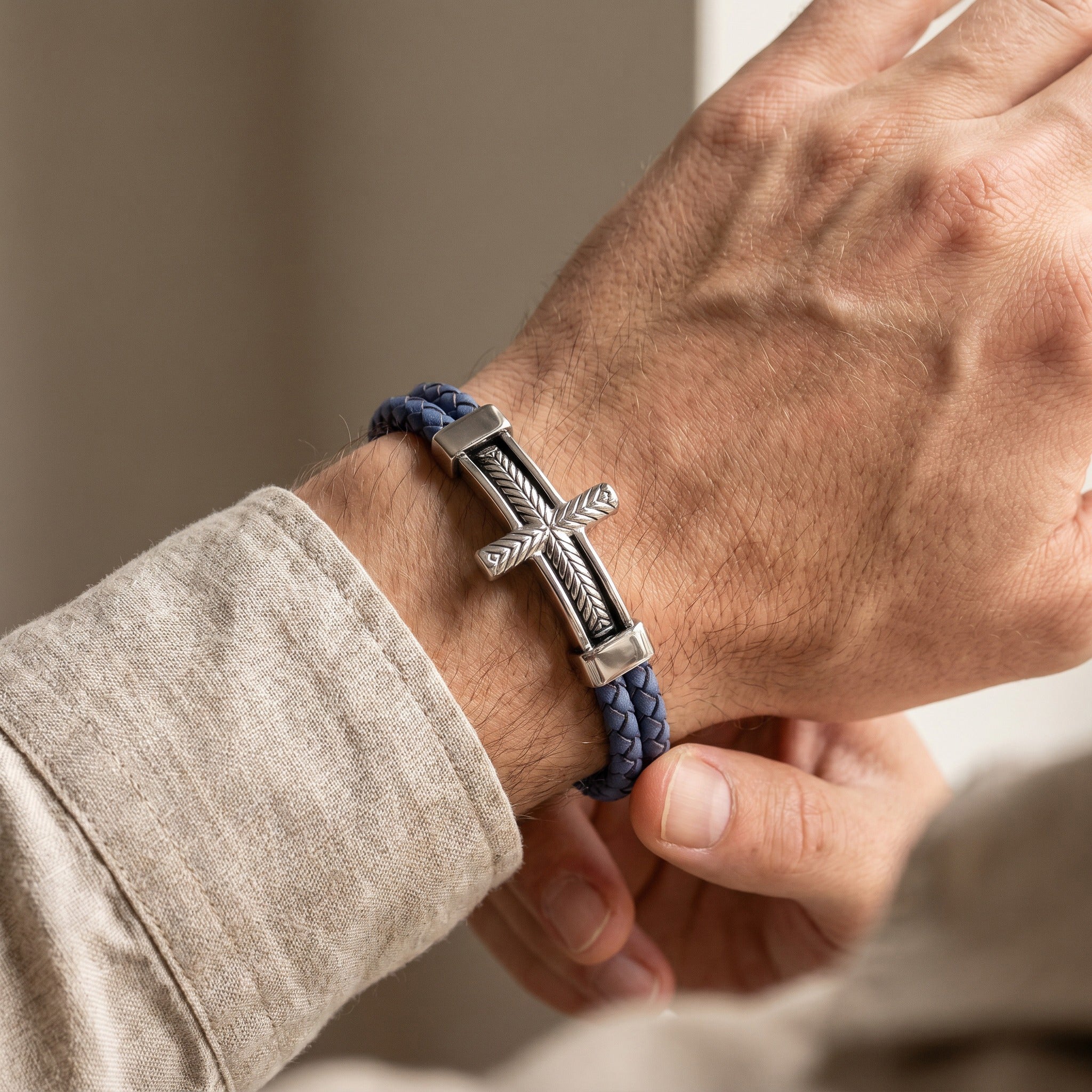 Hand wearing a blue braided bracelet with a silver cross charm on a neutral background