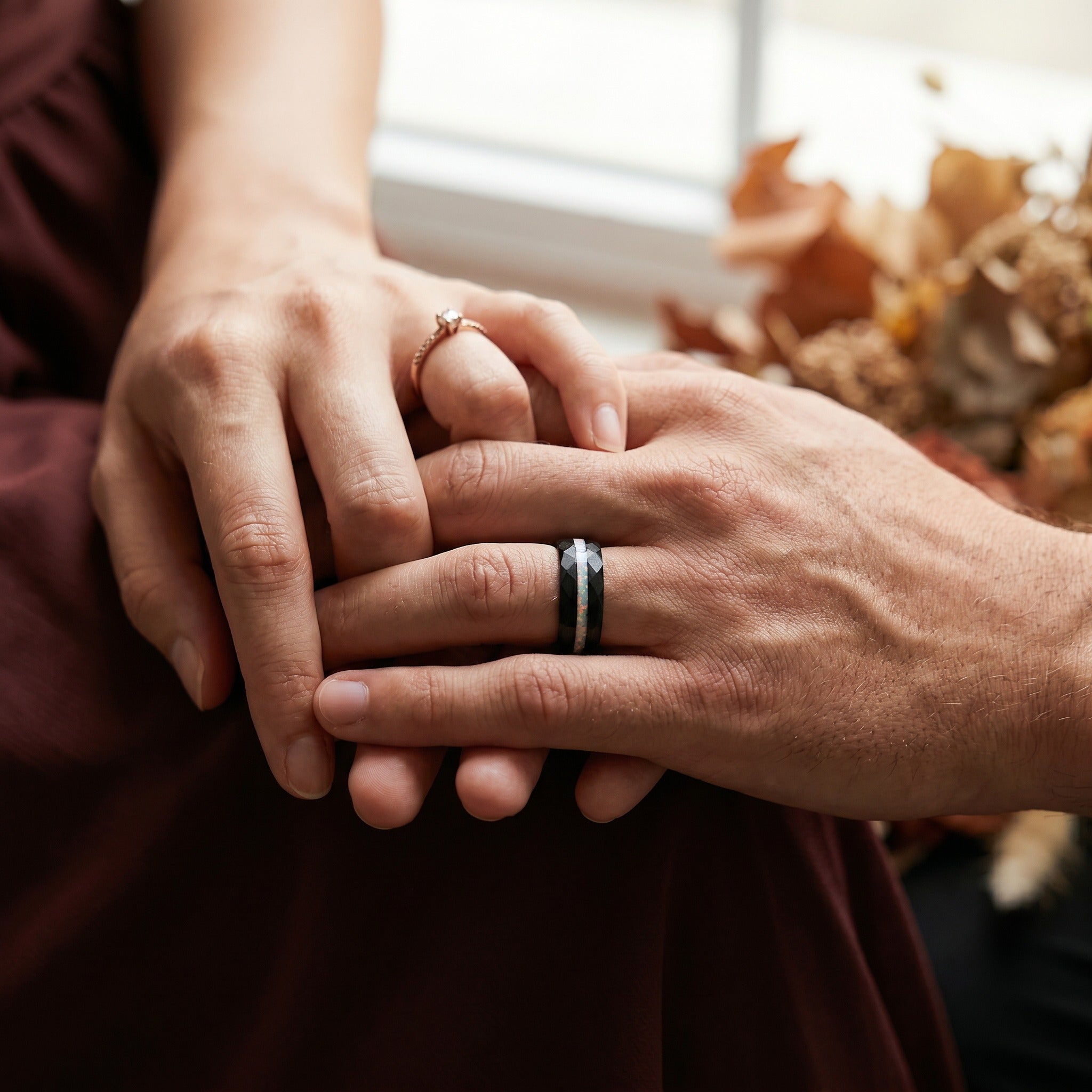 Two hands holding each other with rings on, blurred autumn leaves in the background