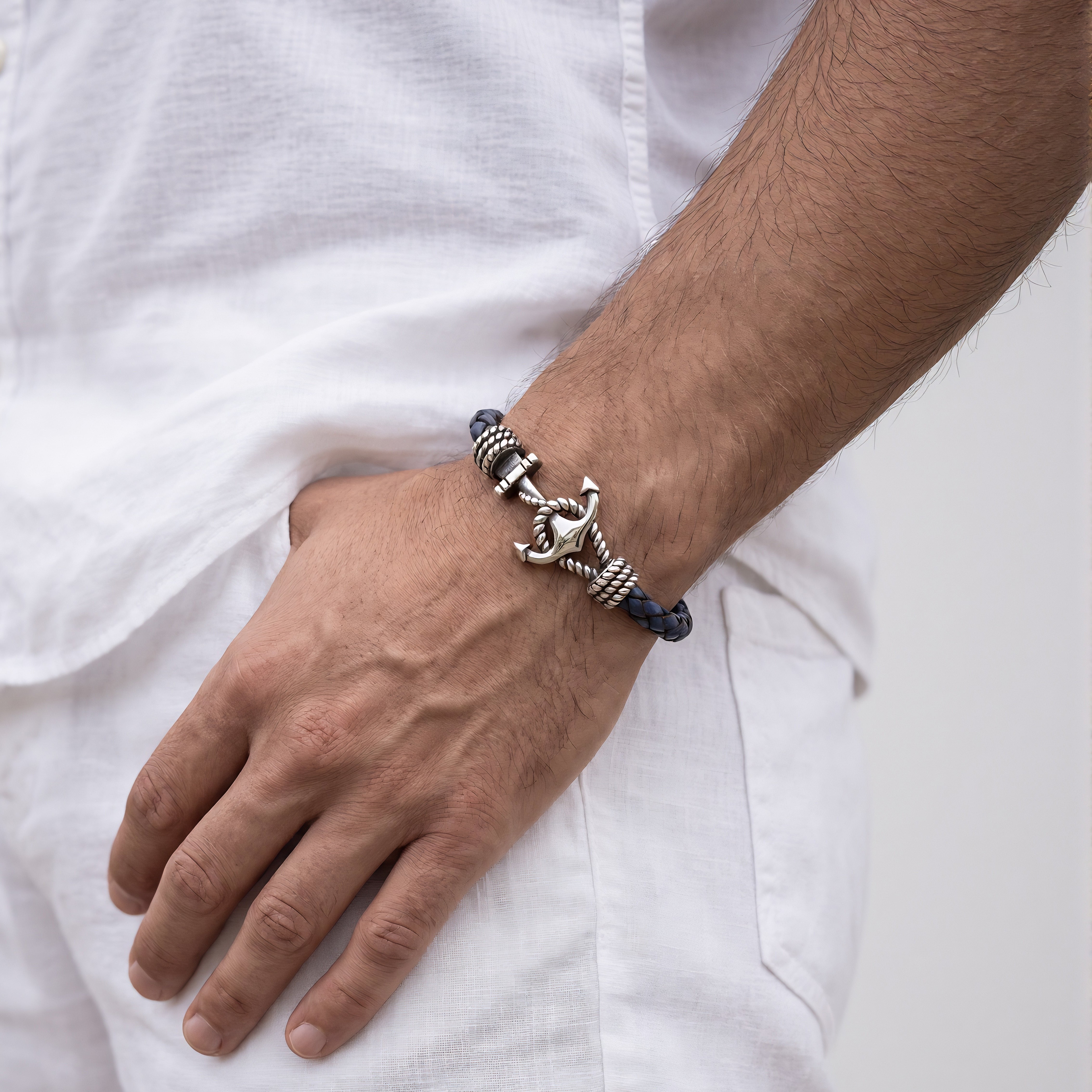 A close-up of a man's wrist wearing a braided navy and silver bracelet with a metallic anchor charm, styled with a white linen shirt.