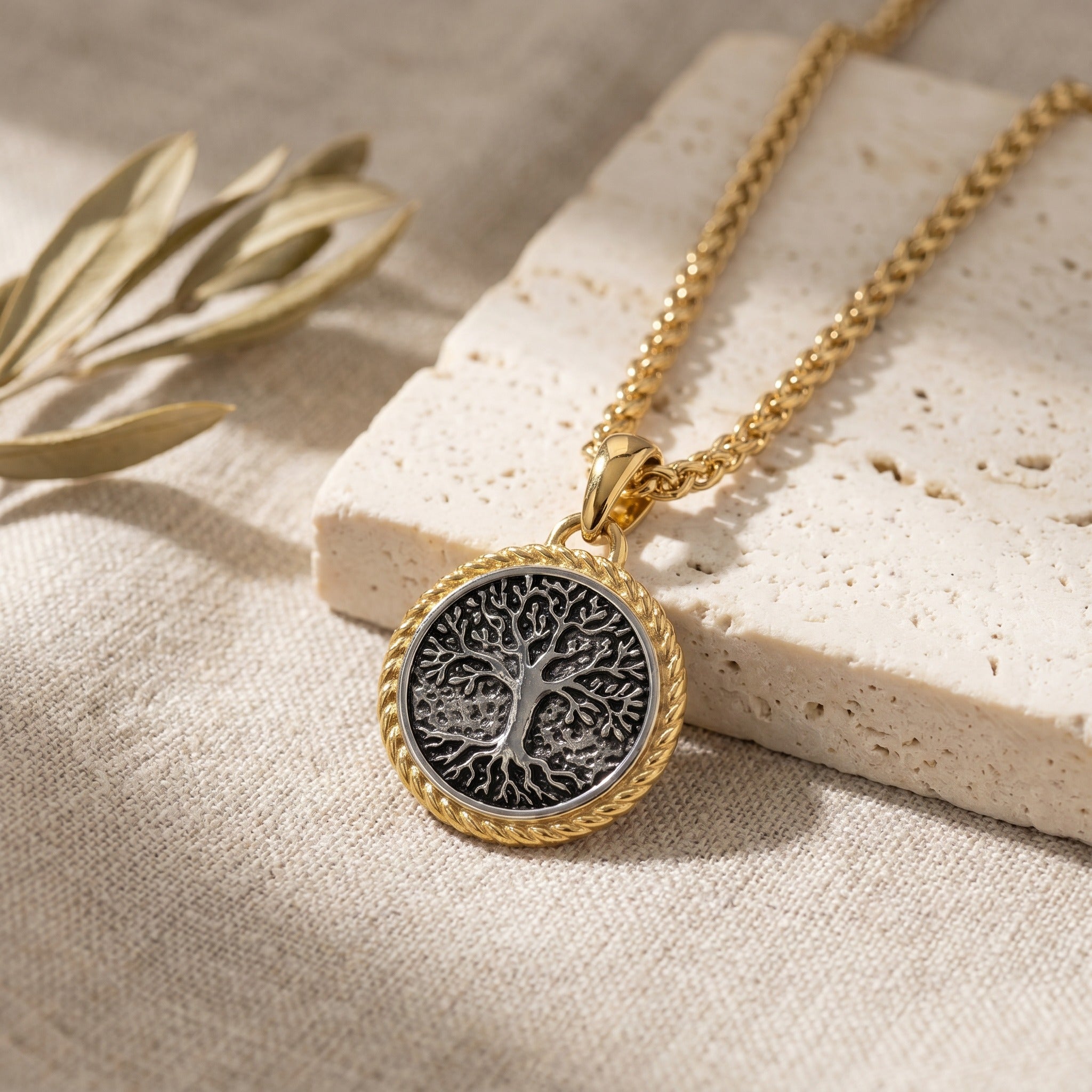 Gold necklace with a tree of life pendant on a textured surface with olive branch