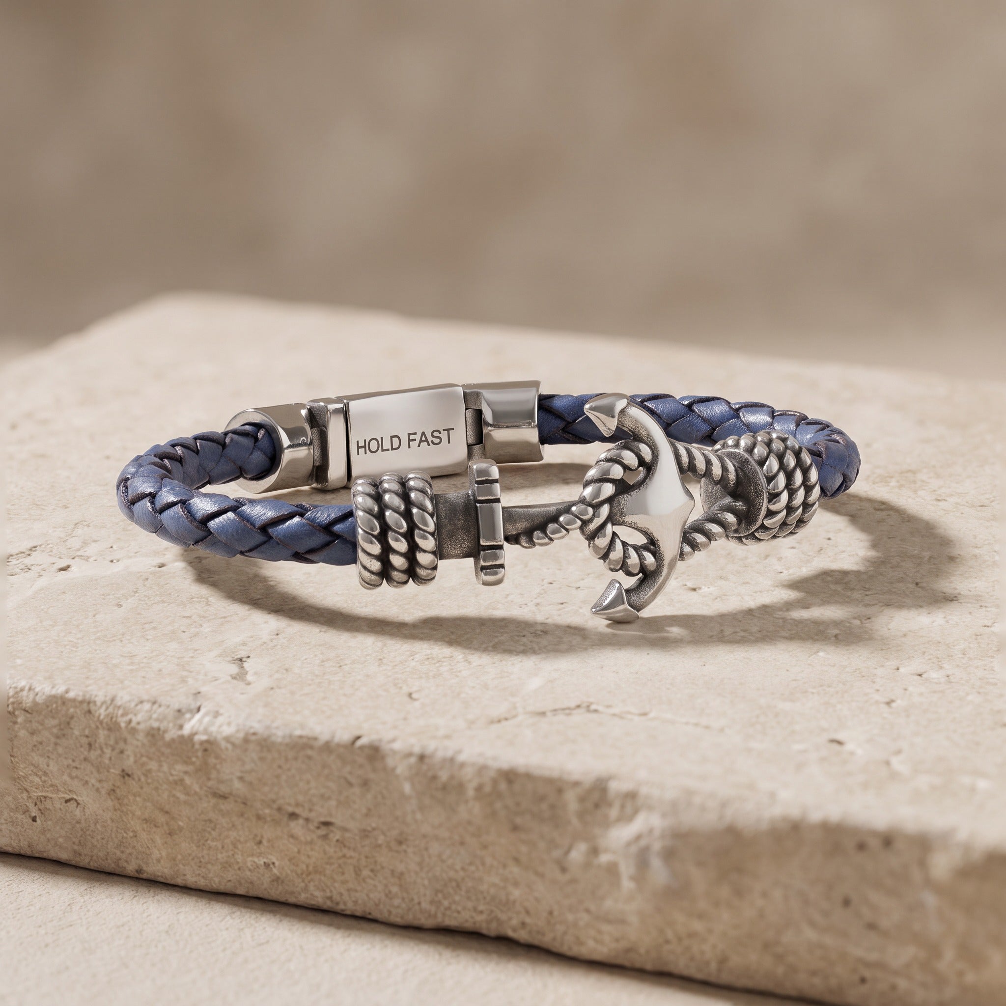 A custom navy blue and silver-toned metal bracelet with an anchor charm and a clasp engraved with the words 'HOLD FAST', displayed on a textured stone slab.