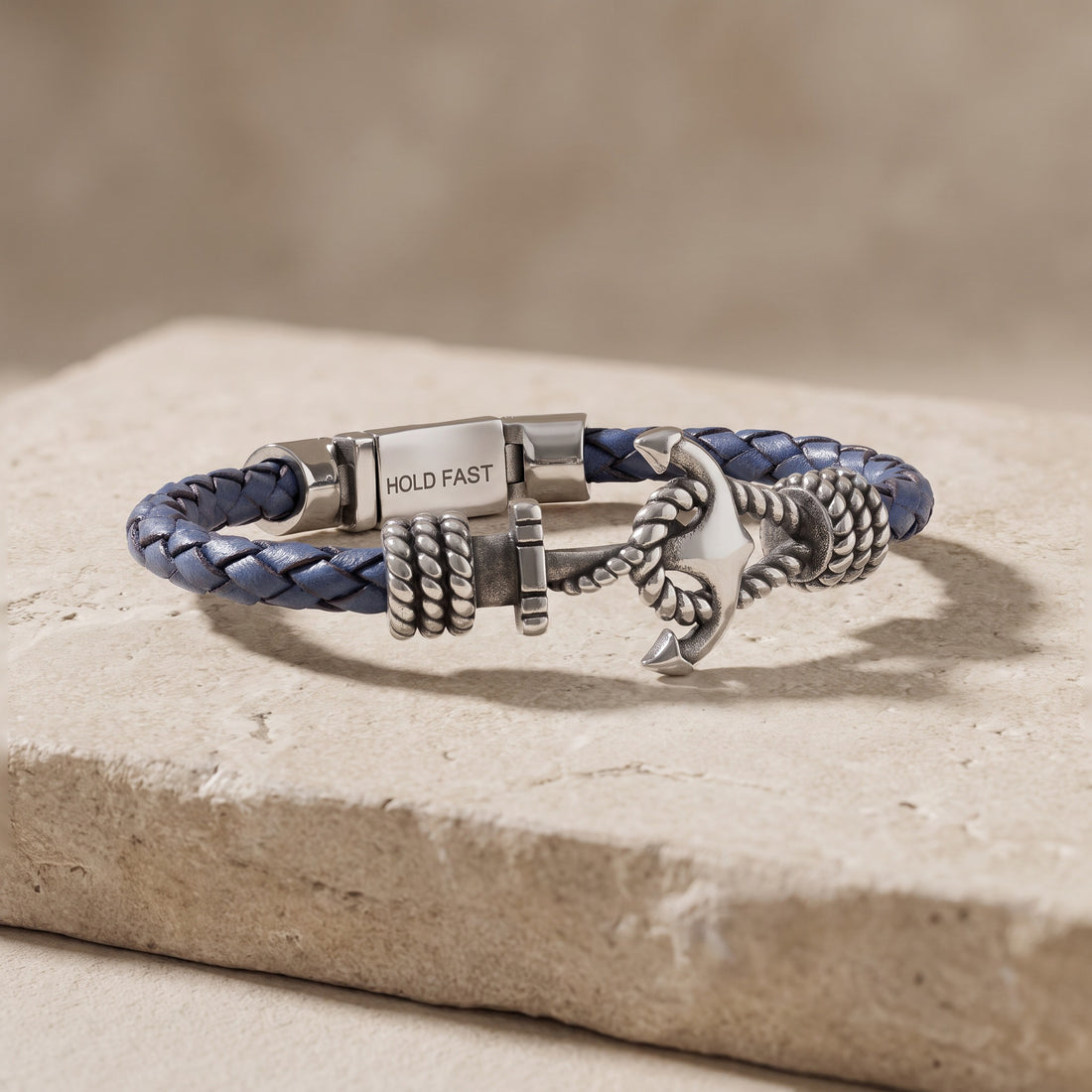 A custom navy blue and silver-toned metal bracelet with an anchor charm and a clasp engraved with the words 'HOLD FAST', displayed on a textured stone slab.