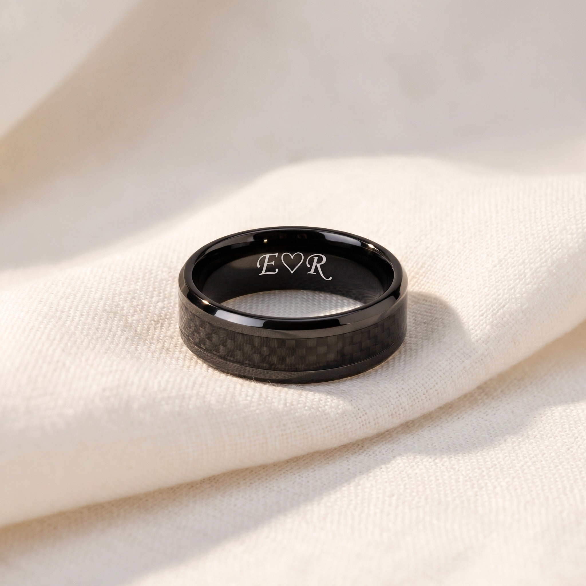 Black carbon fiber ring with engraved initials on a beige fabric background