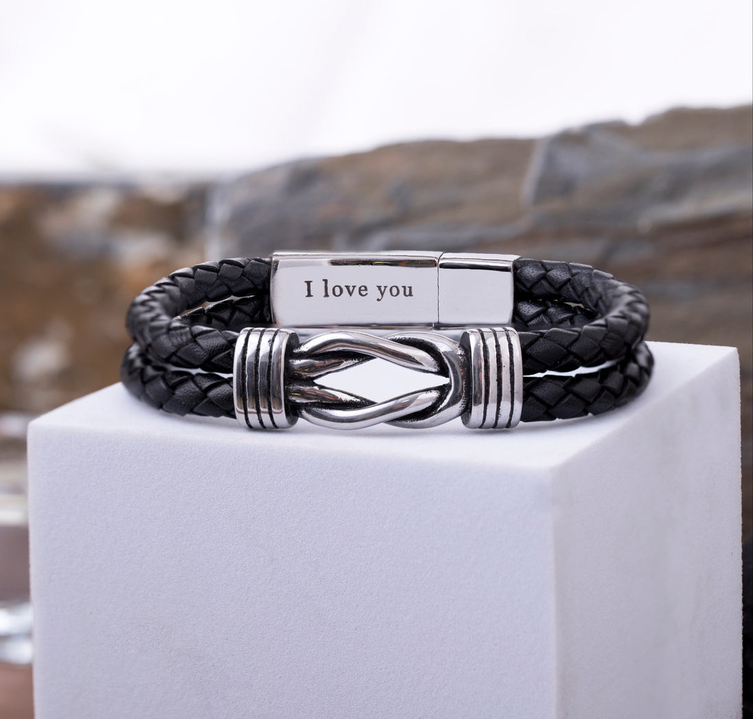 A black braided leather bracelet with a silver-toned metal clasp engraved with 'I love you', displayed on a white cube with a blurred outdoor background.
