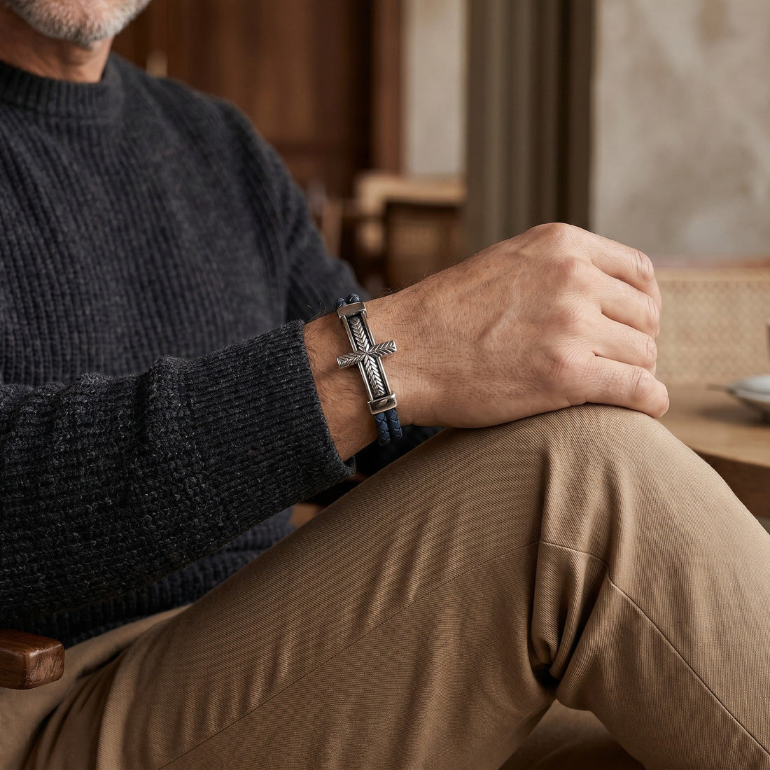 Man wearing a dark sweater and brown pants with a cross bracelet, sitting indoors.
