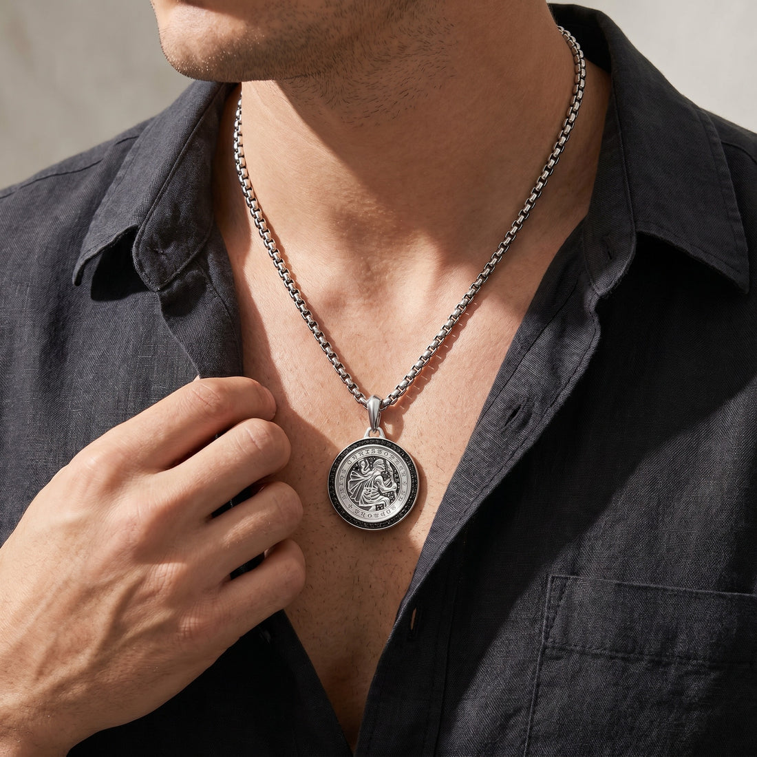 Man wearing a silver necklace with a round pendant, adjusting it with his hand.
