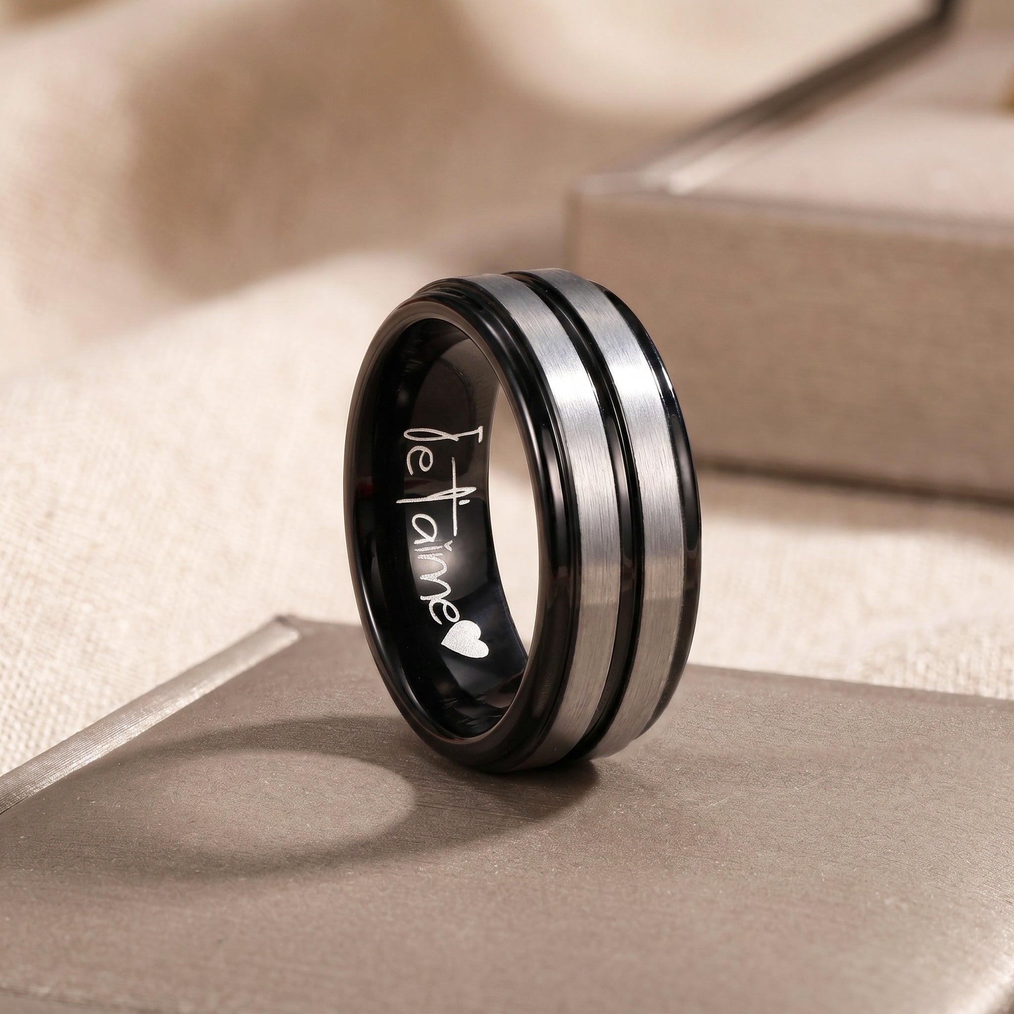 Black and silver ring with 'Je T'aime' engraving on a beige surface