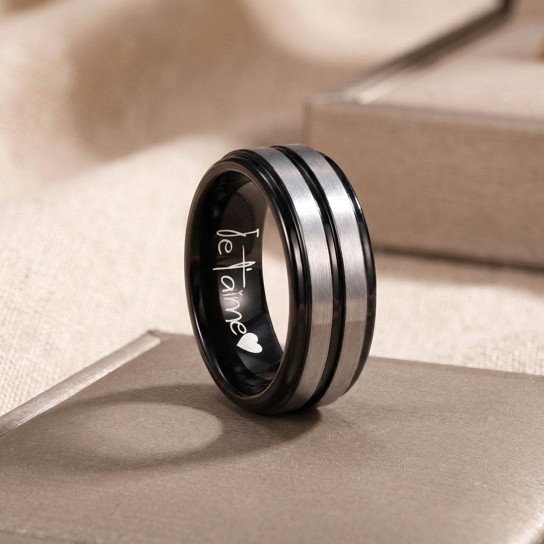 Black and silver ring with 'Je T'aime' engraving on a beige surface